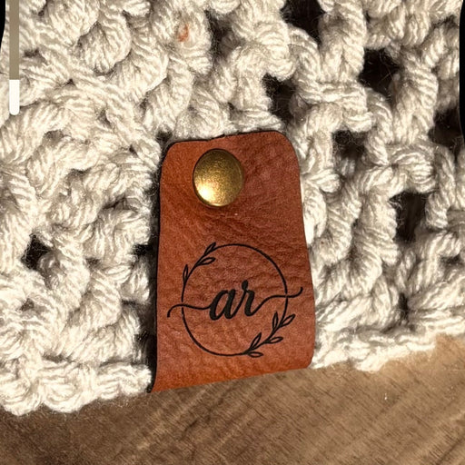 Engraved stylized initials on a leather tag riveted to a crocheted item.