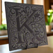 Black granite tile that is engraved around a floral design with the letter K and a solid border.