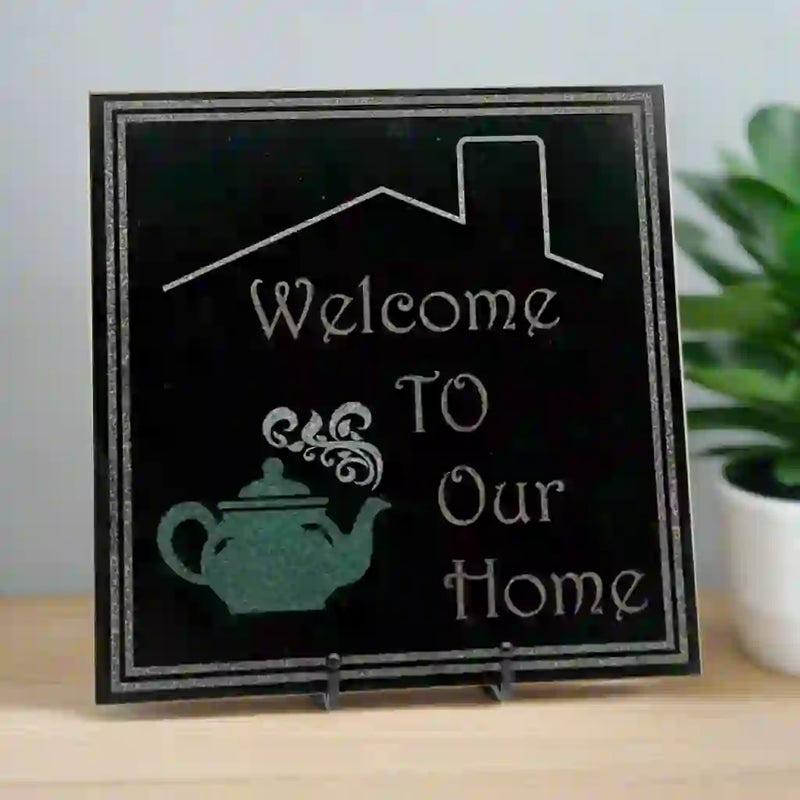 Black granite tile with double box engraving, enclosing 'welcome to our home' text, a teapot on the left, and a house roof above.